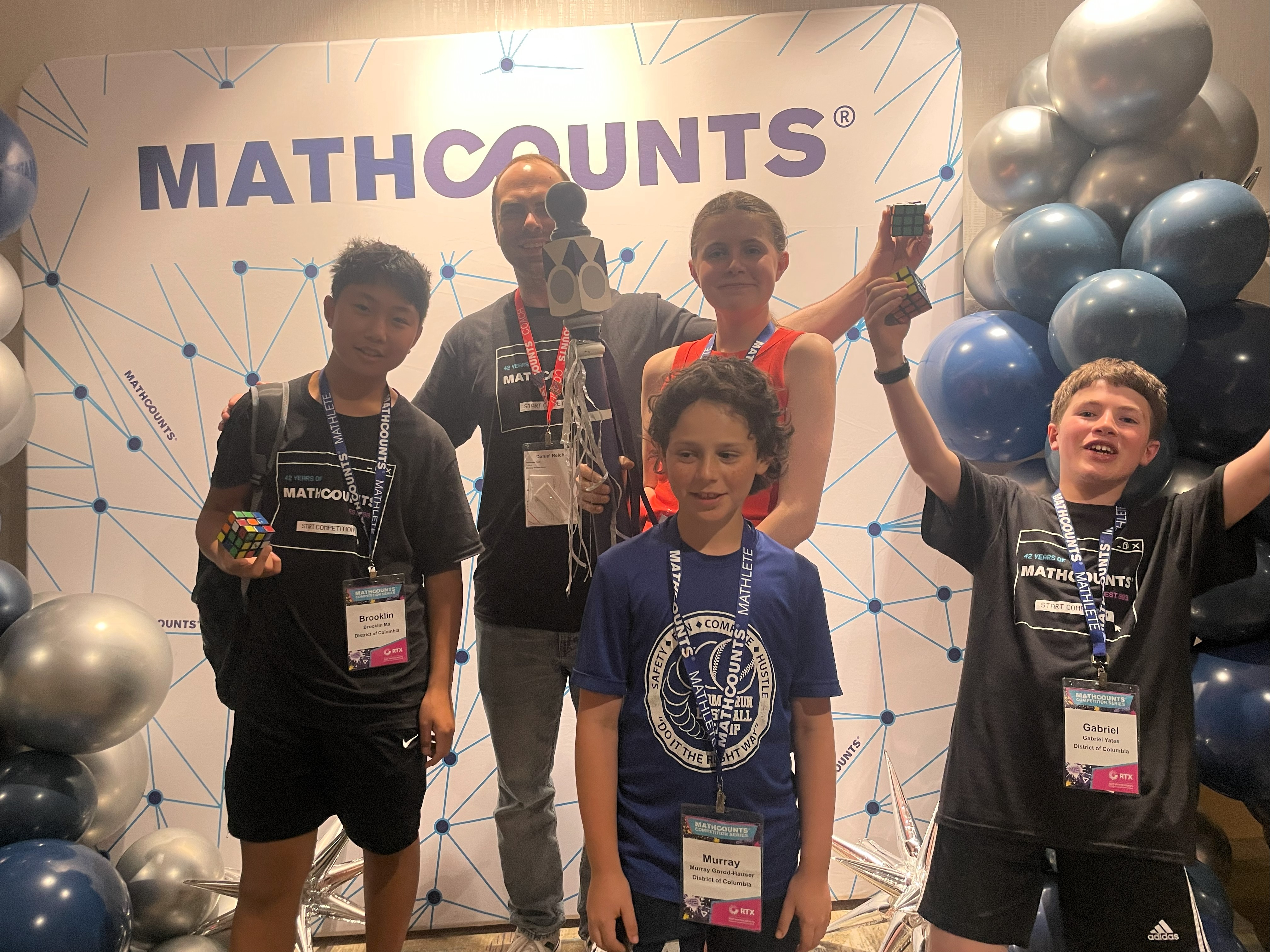 4 students cheer with the spirit stick and their coach in front of a MATHCOUNTS backdrop